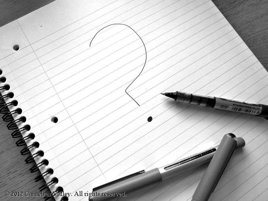 A photograph of an open notepad with a big question mark drawn on it and a pen lying on top